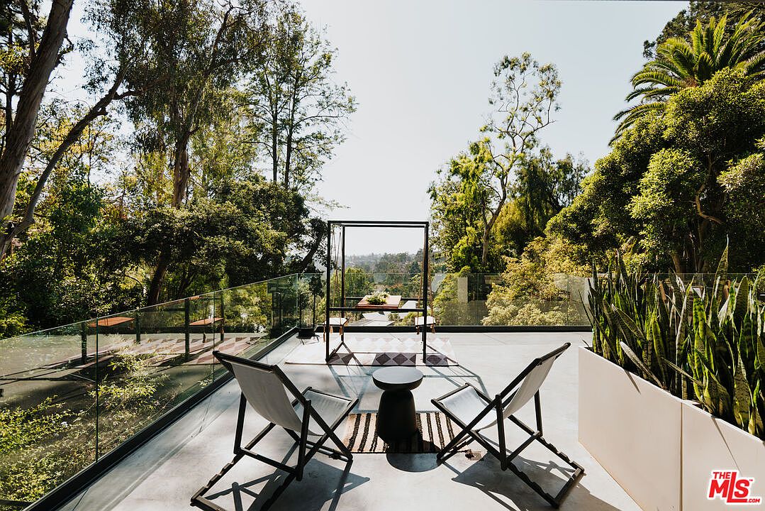 This image showcases a stylish outdoor patio or deck area, featuring modern furniture such as two folding chairs and a small side table arranged on a striped rug. A decorative metal structure frames a dining table in the background, while a glass railing provides unobstructed views of the surrounding lush greenery and trees. The overall impression is one of sophisticated outdoor living and relaxation.