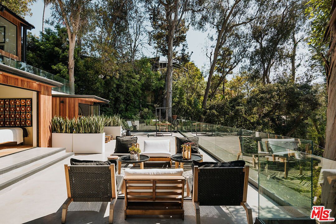 This image showcases a stylish outdoor patio area, featuring modern furniture arranged for comfortable seating. The patio is surrounded by a glass railing, offering unobstructed views of the lush greenery and trees in the background. The design incorporates clean lines and natural materials, creating a serene and inviting outdoor living space.