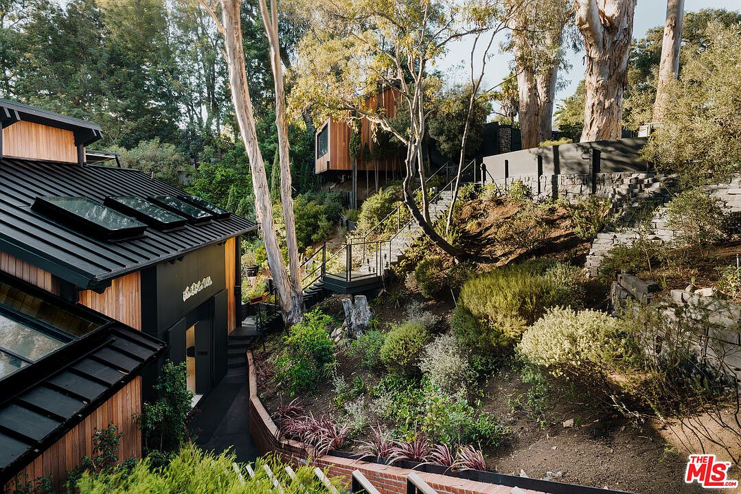 This image showcases a beautifully landscaped yard and garden area. The property features multiple structures, including a modern house with skylights and a unique treehouse-like structure, connected by winding pathways and stairs. Lush greenery, mature trees, and stone retaining walls create a serene and inviting outdoor space.