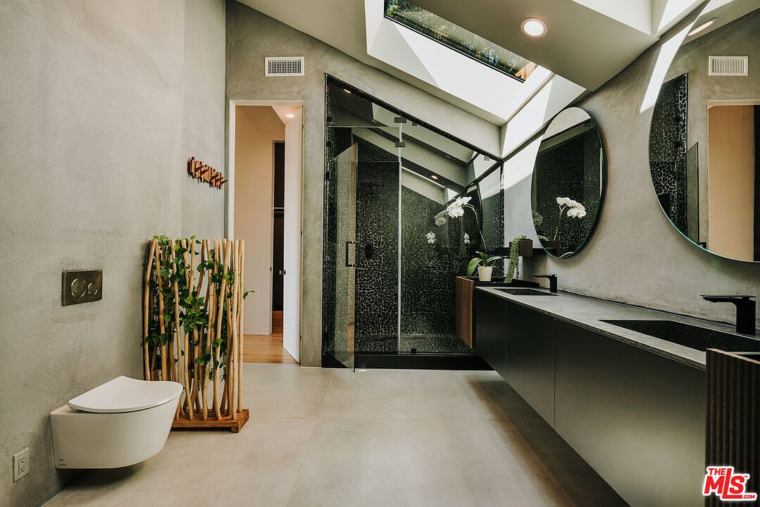 This is a modern primary bathroom featuring a sleek, wall-mounted toilet and a unique wooden screen with greenery. The bathroom boasts a contemporary vanity with dual sinks, illuminated by natural light from a skylight above the shower. The shower is enclosed with glass and has dark, textured walls, adding a touch of luxury.