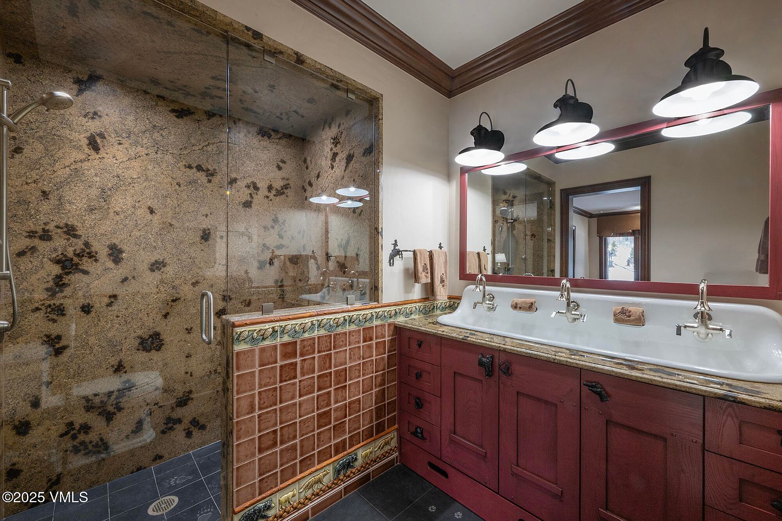 This rustic-style bathroom features a unique trough-style sink with multiple faucets set into a stone countertop atop a deep red wooden vanity. To the left, a walk-in shower is enclosed by glass and finished with textured, earth-toned stone walls and a decorative tile border featuring wildlife motifs. The space is illuminated by three industrial-style pendant lights hanging above a large mirror with a matching red frame.