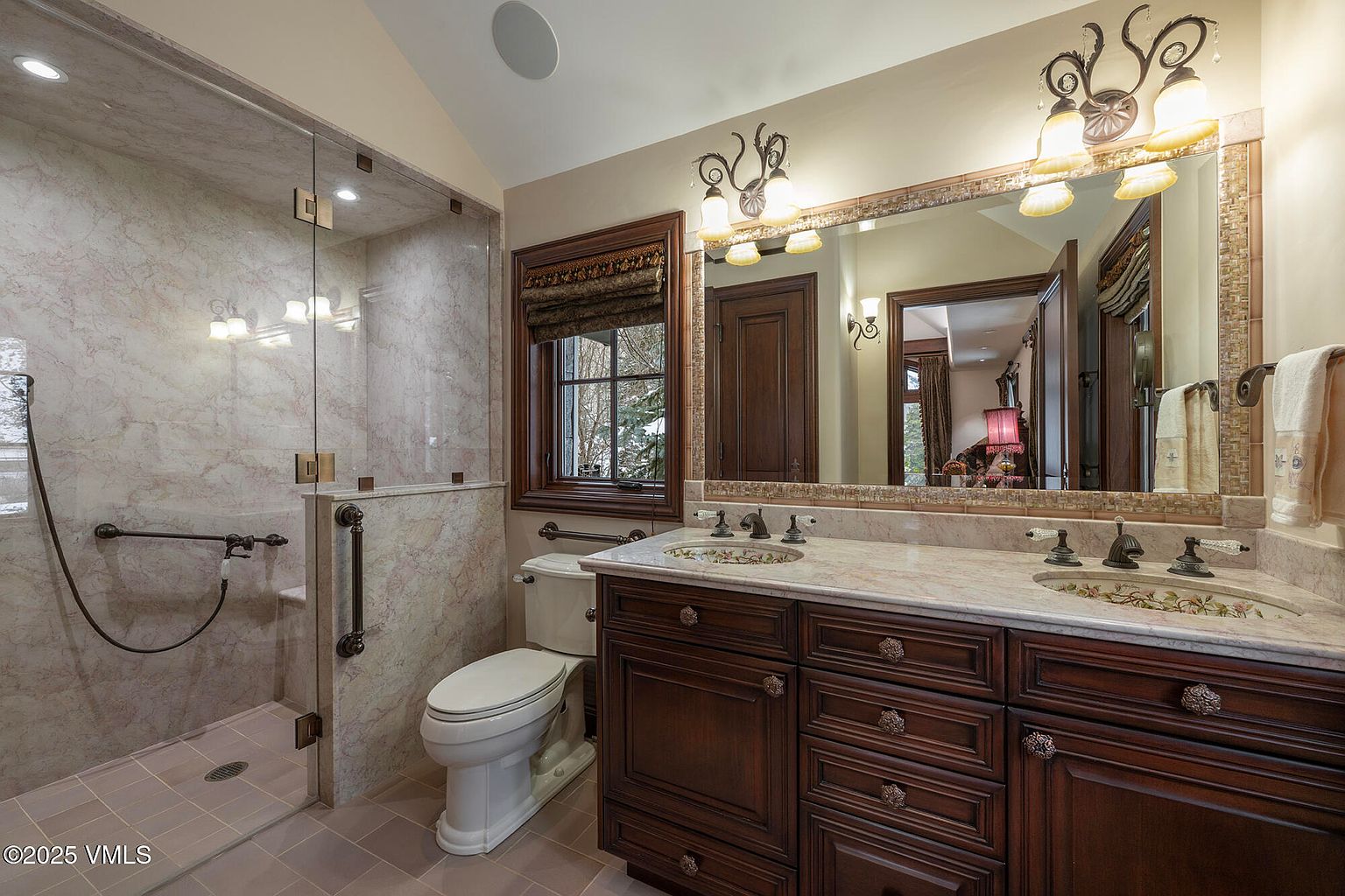 This elegant primary bathroom features a dual-sink vanity with dark wood cabinetry and a light-toned stone countertop. A large, glass-enclosed walk-in shower with patterned stone walls sits adjacent to a white toilet and a window framed in dark wood. The space is illuminated by decorative wall-mounted light fixtures and a large mirror that reflects the adjacent room.