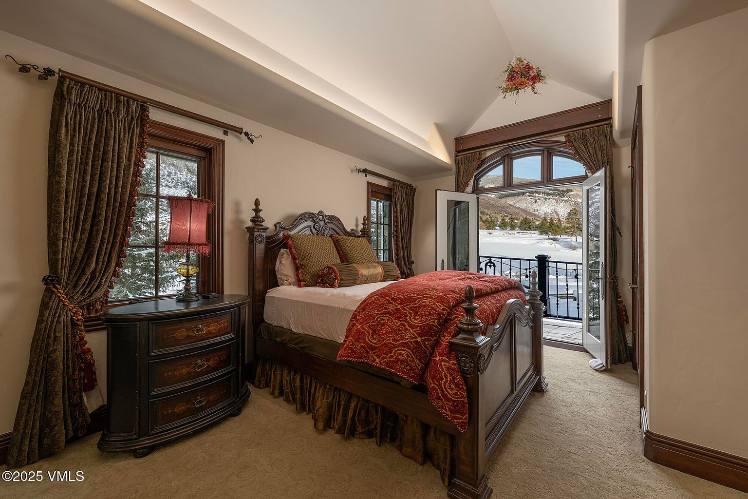 This elegant bedroom features a traditional wooden bed frame with a rich red patterned comforter and decorative pillows. A dark wood dresser with a matching lamp sits to the left, while a set of French doors opens onto a balcony with a scenic, snow-covered mountain view. The room is accented by warm lighting, heavy drapery, and a vaulted ceiling, creating a cozy and sophisticated atmosphere.