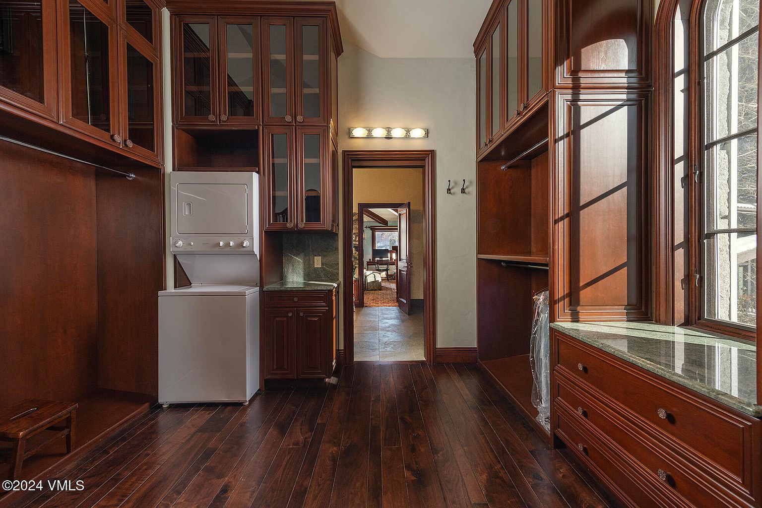 This elegant laundry room features rich, dark wood cabinetry with glass-front upper cabinets and integrated storage solutions. A stacked washer and dryer unit is positioned next to a stone-topped counter, while a large window on the right allows natural light to illuminate the dark wood flooring. The space serves as a functional transition area, with a doorway leading into an adjacent room, creating a sophisticated and organized utility space.