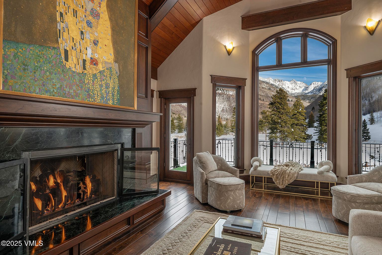 This elegant living room features a prominent fireplace with a dark stone surround and a large, iconic art piece mounted above the mantel. The space is anchored by a comfortable seating area with plush armchairs and a bench, all oriented toward expansive windows that frame a breathtaking view of snow-capped mountains. The room is finished with rich wood flooring and a vaulted wood-paneled ceiling, creating a warm and sophisticated mountain retreat atmosphere.