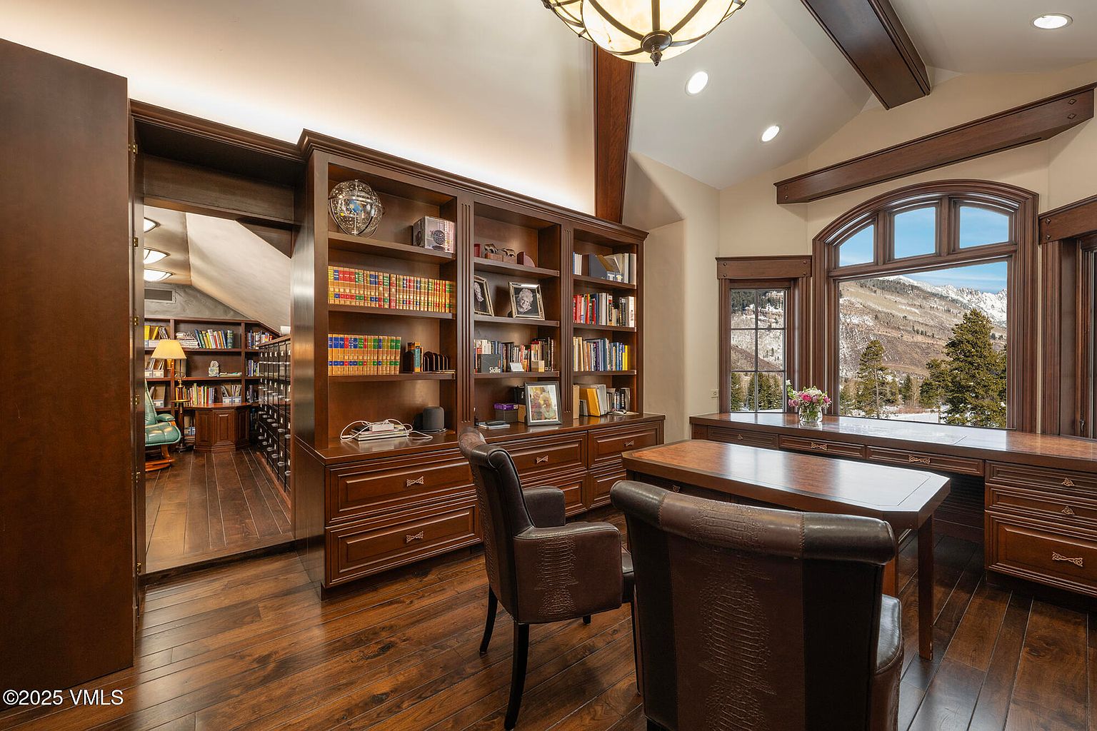 This sophisticated home office features extensive dark wood built-in cabinetry and shelving that spans the wall, providing ample storage and display space. A large desk sits in the center of the room, complemented by leather chairs, while a wide window offers a scenic view of snow-capped mountains. The room is connected to an adjacent library or study area, creating a cohesive and professional atmosphere with warm lighting and rich wood tones.