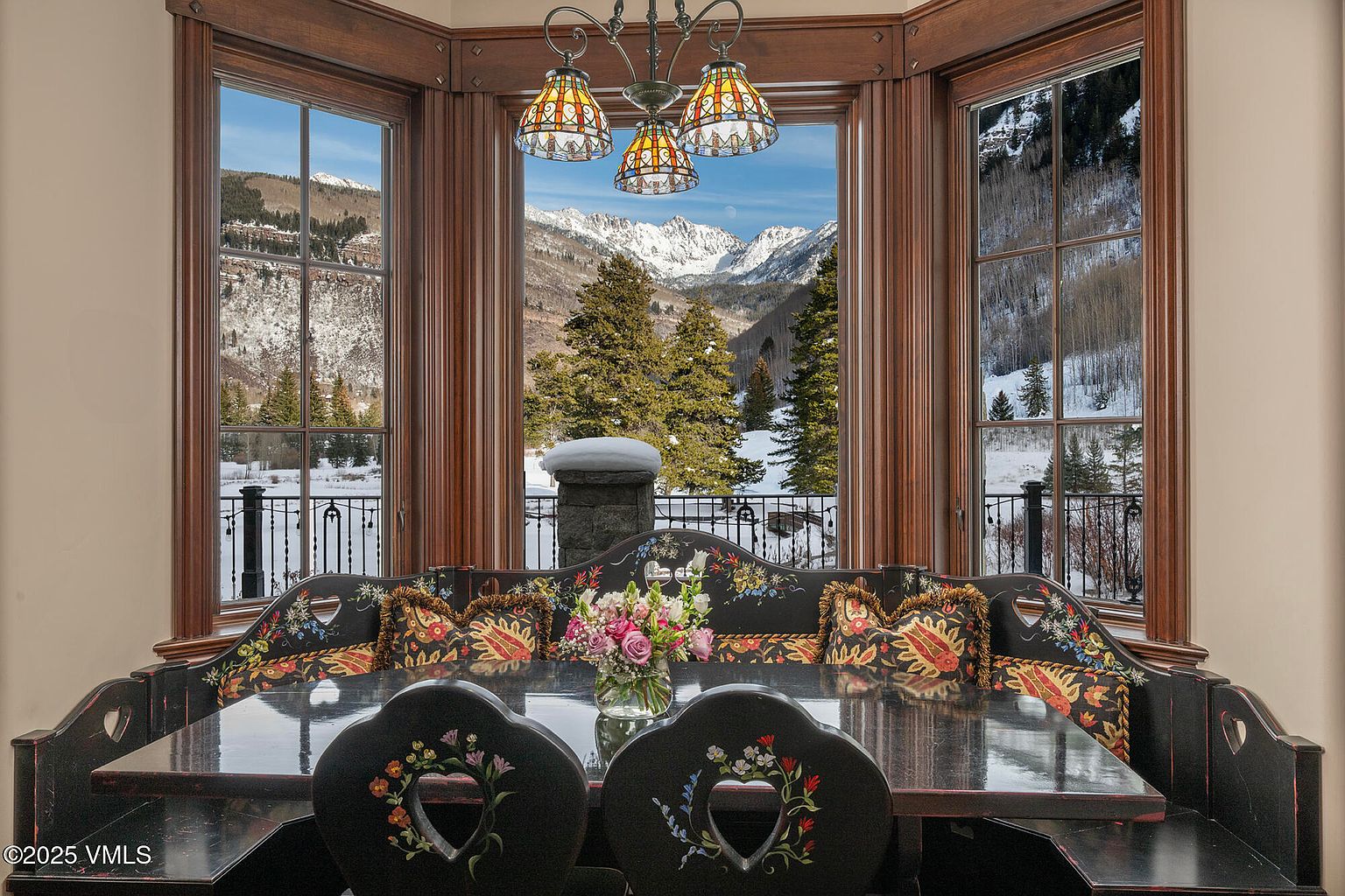 This dining area features a rustic, hand-painted wooden bench and matching chairs set against a large bay window that frames a breathtaking mountain view. The space is illuminated by a trio of ornate, stained-glass pendant lights hanging above a dark, reflective dining table adorned with a fresh floral centerpiece. The combination of traditional folk-art furniture and the dramatic, snow-covered alpine backdrop creates a cozy, high-end mountain retreat atmosphere.
