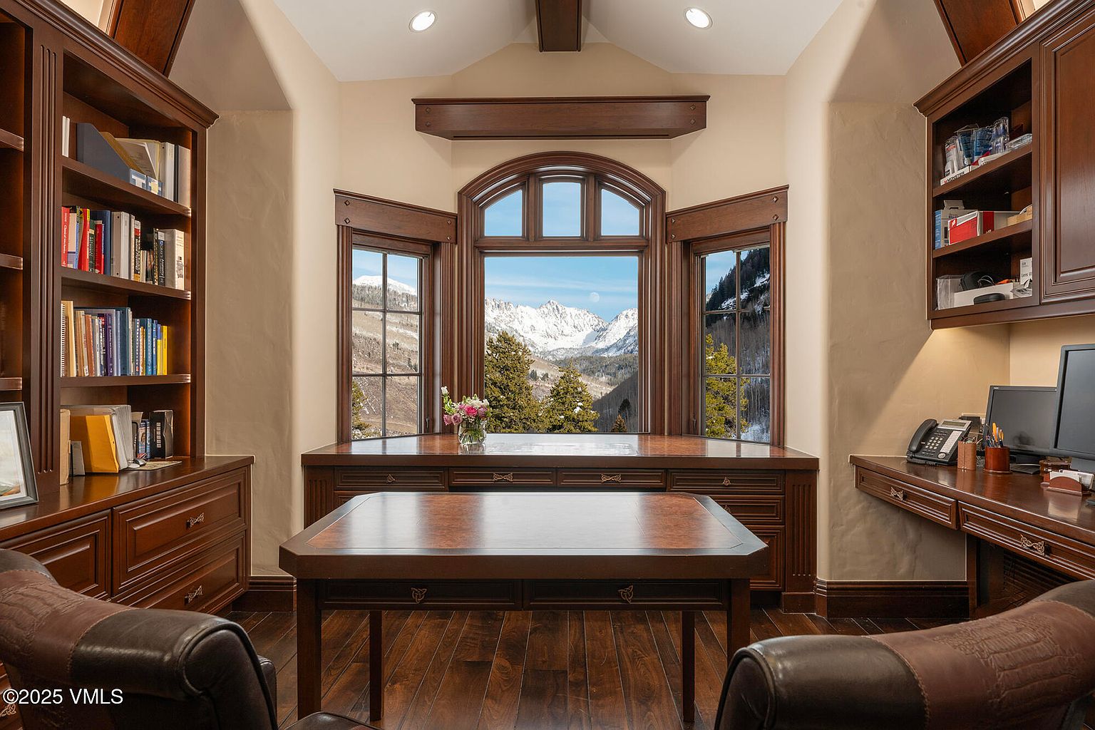 This sophisticated home office features rich, dark wood built-in cabinetry and shelving that frame a central bay window with breathtaking mountain views. A large, matching wood desk sits in the foreground, creating a professional and focused workspace. The room is accented by warm lighting and leather seating, offering a serene and luxurious atmosphere perfect for productivity.