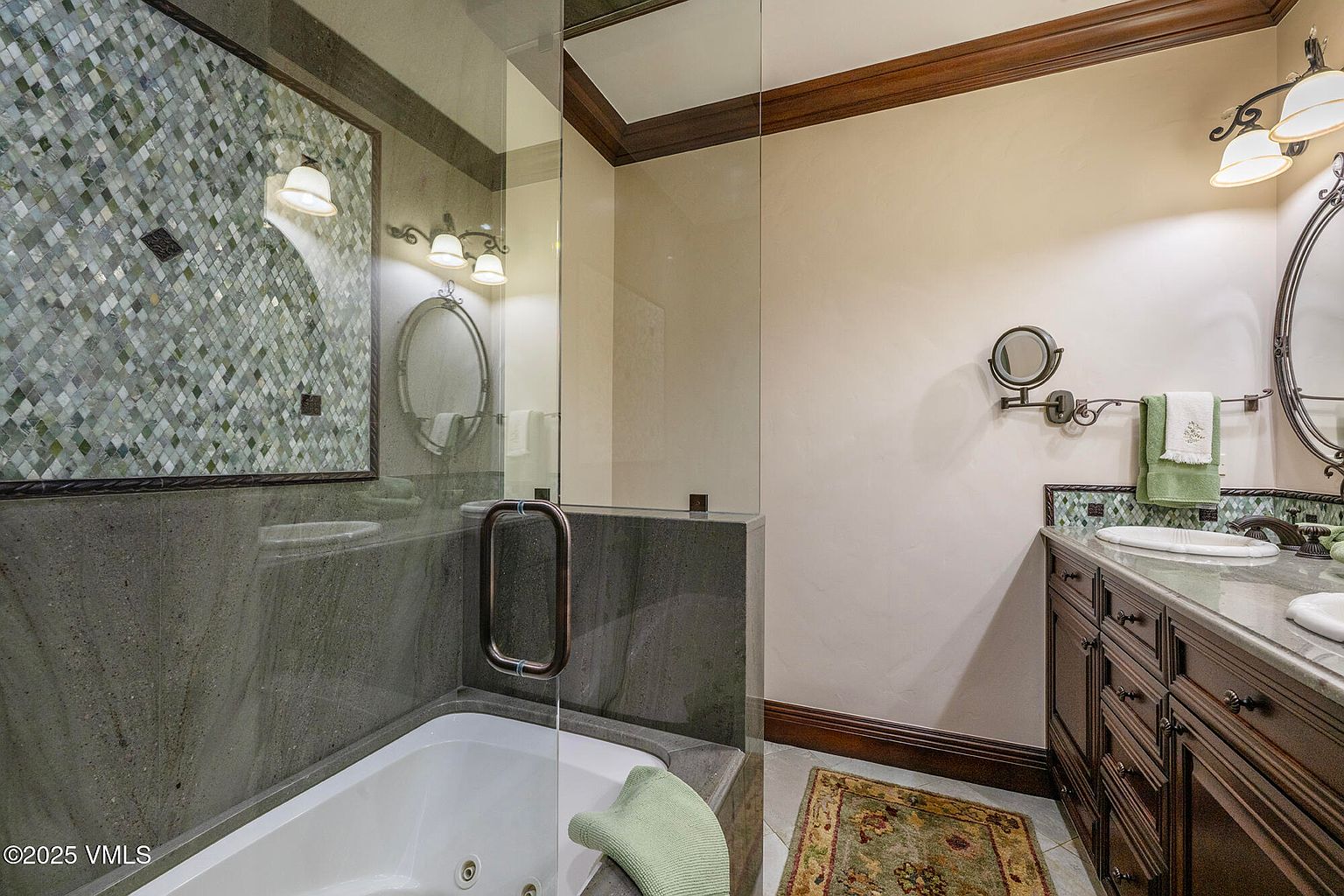 This elegant bathroom features a deep soaking tub enclosed by a glass partition and framed by dark stone walls and a decorative mosaic tile accent. To the right, a dark wood vanity with a stone countertop and an oval mirror creates a sophisticated, traditional aesthetic. The space is illuminated by warm wall-mounted lighting and includes a patterned area rug, contributing to a luxurious and cozy atmosphere.