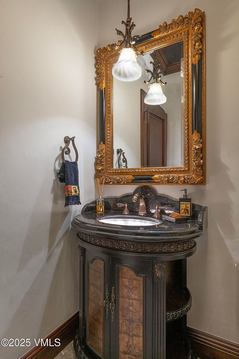 This elegant powder room features a dark, ornate vanity with a stone countertop and an integrated sink. Above the vanity hangs a large, intricately carved gold-toned mirror, complemented by a decorative hanging light fixture. The space is finished with a wall-mounted towel ring and a soap dispenser, creating a sophisticated and traditional aesthetic.