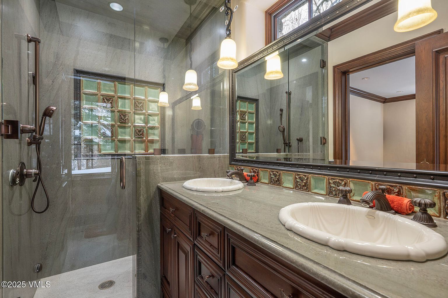 This primary bathroom features a dual-sink vanity with dark wood cabinetry and a stone countertop, complemented by a large mirror framed in dark wood. A glass-enclosed shower area is integrated into the space, showcasing stone wall tiles and a decorative accent band of green glass tiles that continues along the vanity backsplash. The room is illuminated by warm pendant lighting, creating a sophisticated and rustic atmosphere.