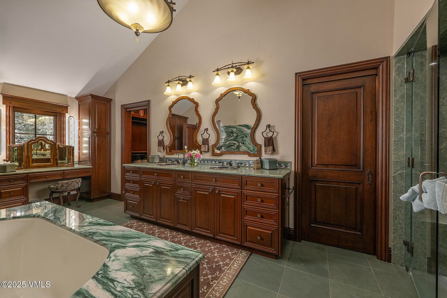This spacious primary bathroom features a double vanity with dark wood cabinetry and a green stone countertop, complemented by two ornate mirrors and wall-mounted light fixtures. To the left, a dedicated vanity area with a makeup mirror and chair is positioned near a window, while a large soaking tub with a matching stone surround occupies the foreground. The room is finished with dark tile flooring and a tall, vaulted ceiling, creating an elegant and traditional atmosphere.