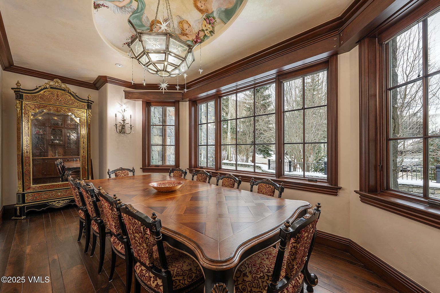This elegant dining room features a large, dark wood table surrounded by ornate chairs with patterned upholstery. The space is defined by rich wood trim, a decorative ceiling mural, and a large, multi-tiered chandelier that serves as a focal point. A tall, decorative cabinet with gold detailing stands against the wall, while large windows provide a view of the snowy exterior.