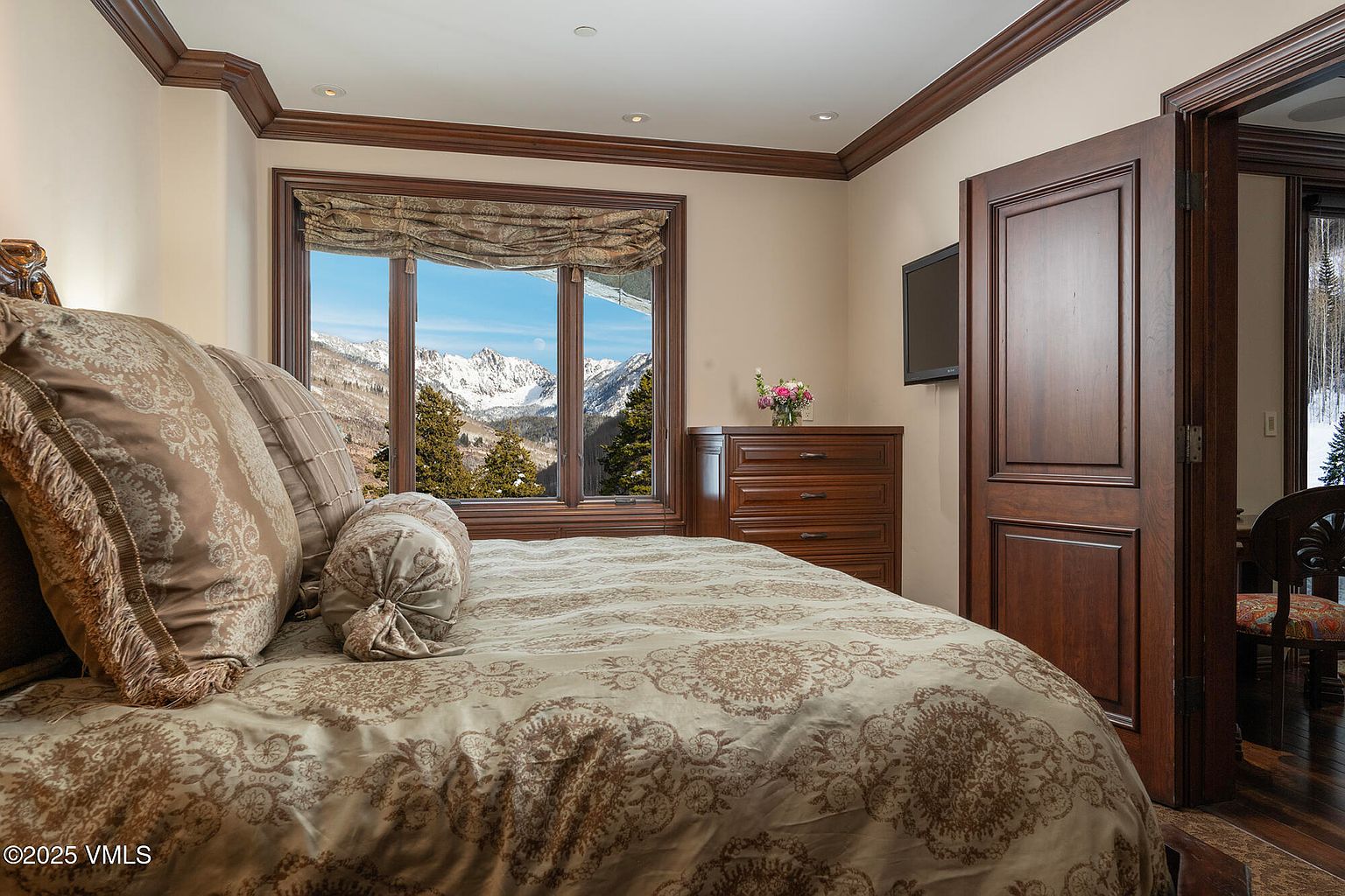 This cozy bedroom features a plush bed with patterned bedding and a decorative bolster pillow, positioned to face a large window with a stunning mountain view. To the right, a dark wood dresser sits beneath a wall-mounted television, while a doorway leads into an adjacent room. The room is accented with crown molding and warm, neutral wall tones, creating a serene and inviting atmosphere.
