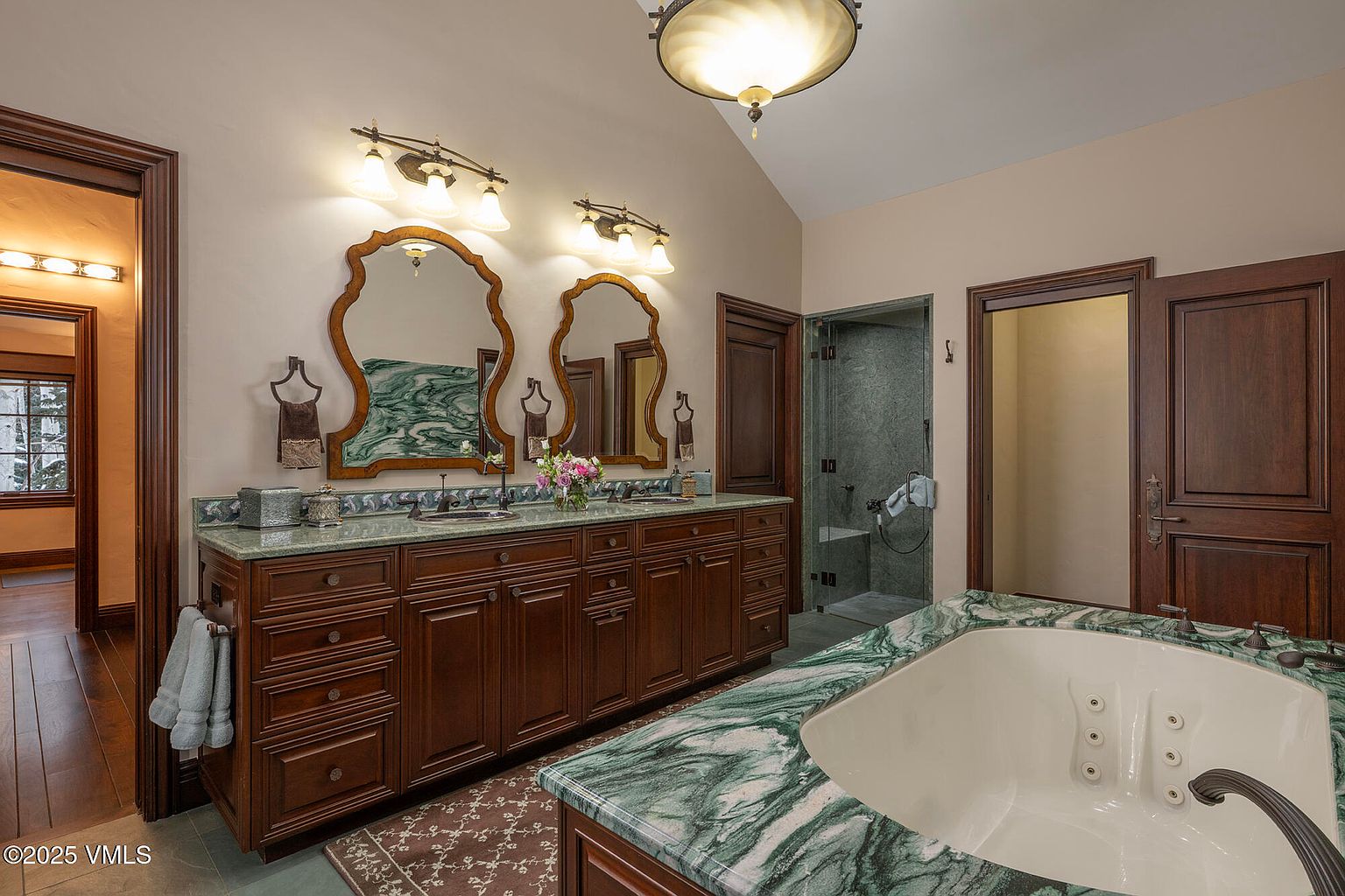 This primary bathroom features a double vanity with dark wood cabinetry and a green-veined stone countertop, complemented by two ornate mirrors and wall-mounted lighting. A large soaking tub with matching stone surround occupies the foreground, while a glass-enclosed shower stall is visible in the background. The space is finished with warm-toned walls and dark wood trim, creating a traditional and luxurious atmosphere.