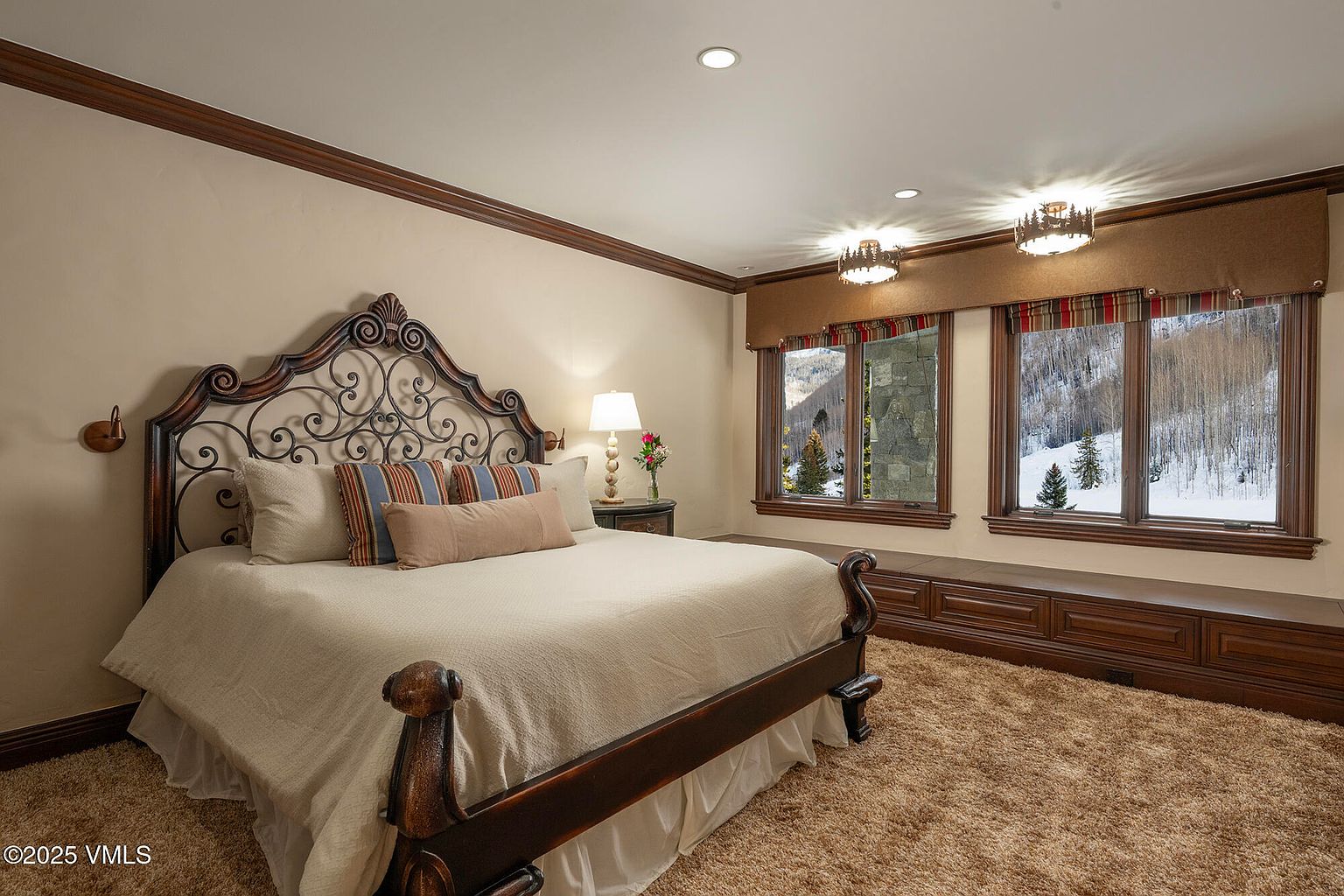 This primary bedroom features a large bed with an ornate, dark-toned metal headboard as the central focal point. The room is carpeted in a neutral tone and includes a built-in window seat beneath two large windows that offer a scenic view of a snowy, wooded landscape. The space is accented by warm, recessed lighting and a bedside lamp, creating a cozy and inviting atmosphere.