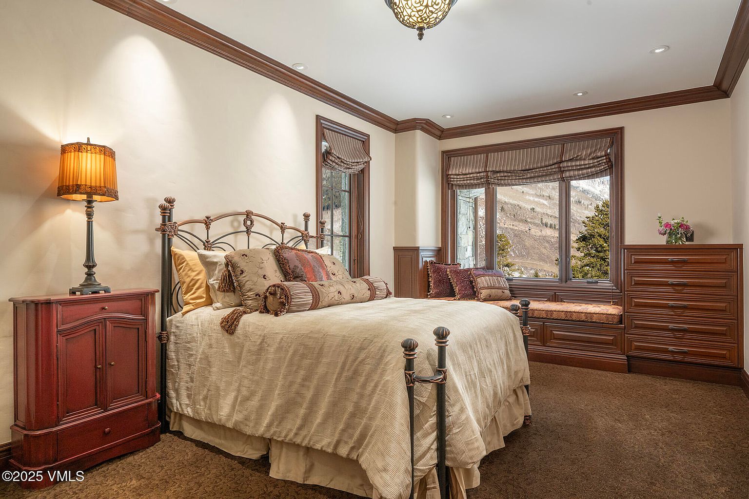 This cozy bedroom features a traditional metal-framed bed dressed in neutral linens, flanked by a dark wood dresser and a matching nightstand with a warm-toned lamp. A built-in window seat with plush cushions sits beneath large windows that offer a scenic view of the mountainous landscape. The room is finished with rich wood trim, crown molding, and wall-to-wall carpeting, creating a warm and inviting atmosphere.