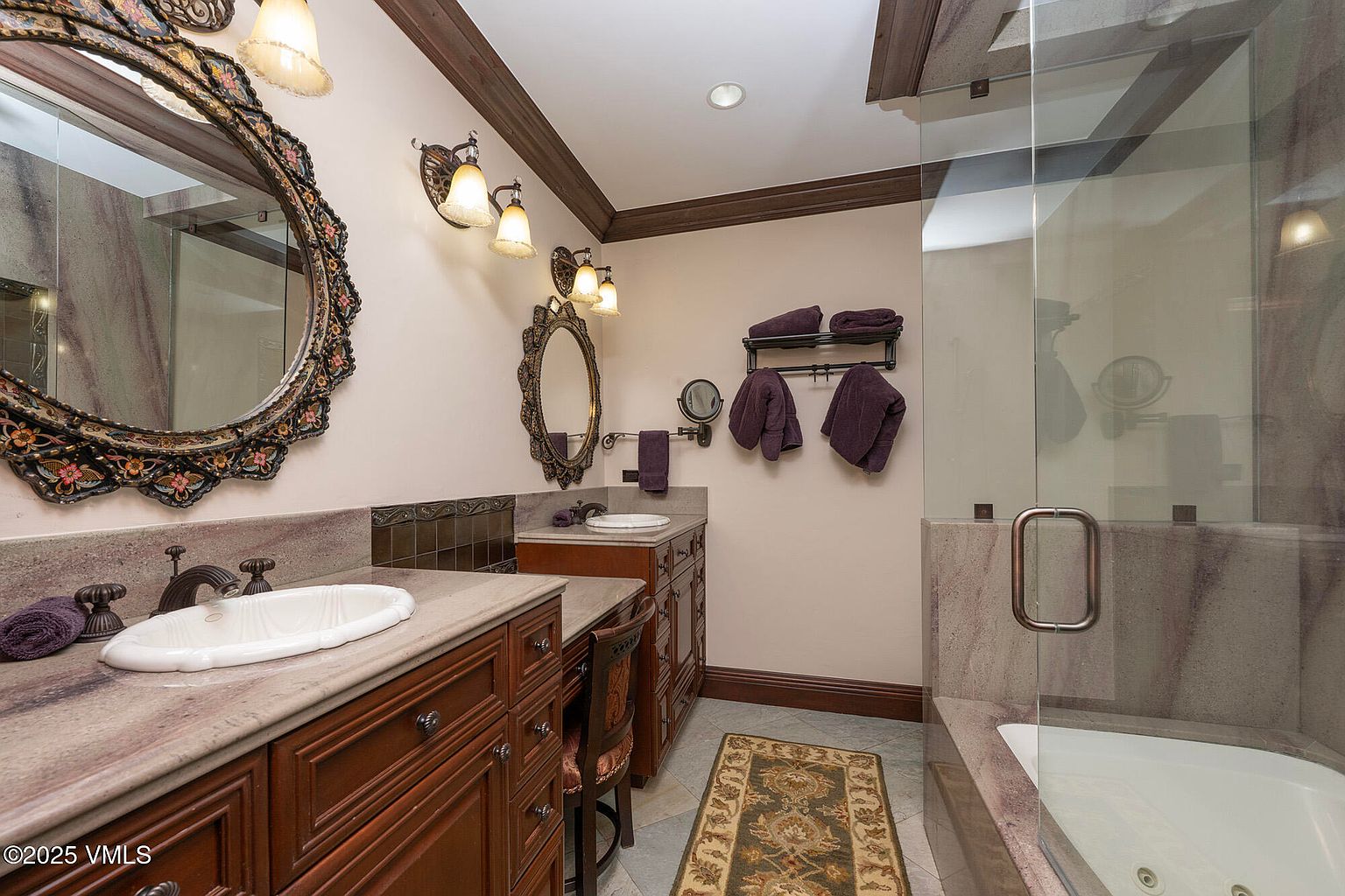 This primary bathroom features a dual-vanity setup with rich-toned wood cabinetry and stone countertops, complemented by ornate, dark-framed mirrors. A glass-enclosed shower area with a built-in tub is positioned to the right, while a patterned rug adds texture to the tiled floor. The space is illuminated by warm wall-mounted lighting and recessed ceiling lights, creating a sophisticated and traditional atmosphere.