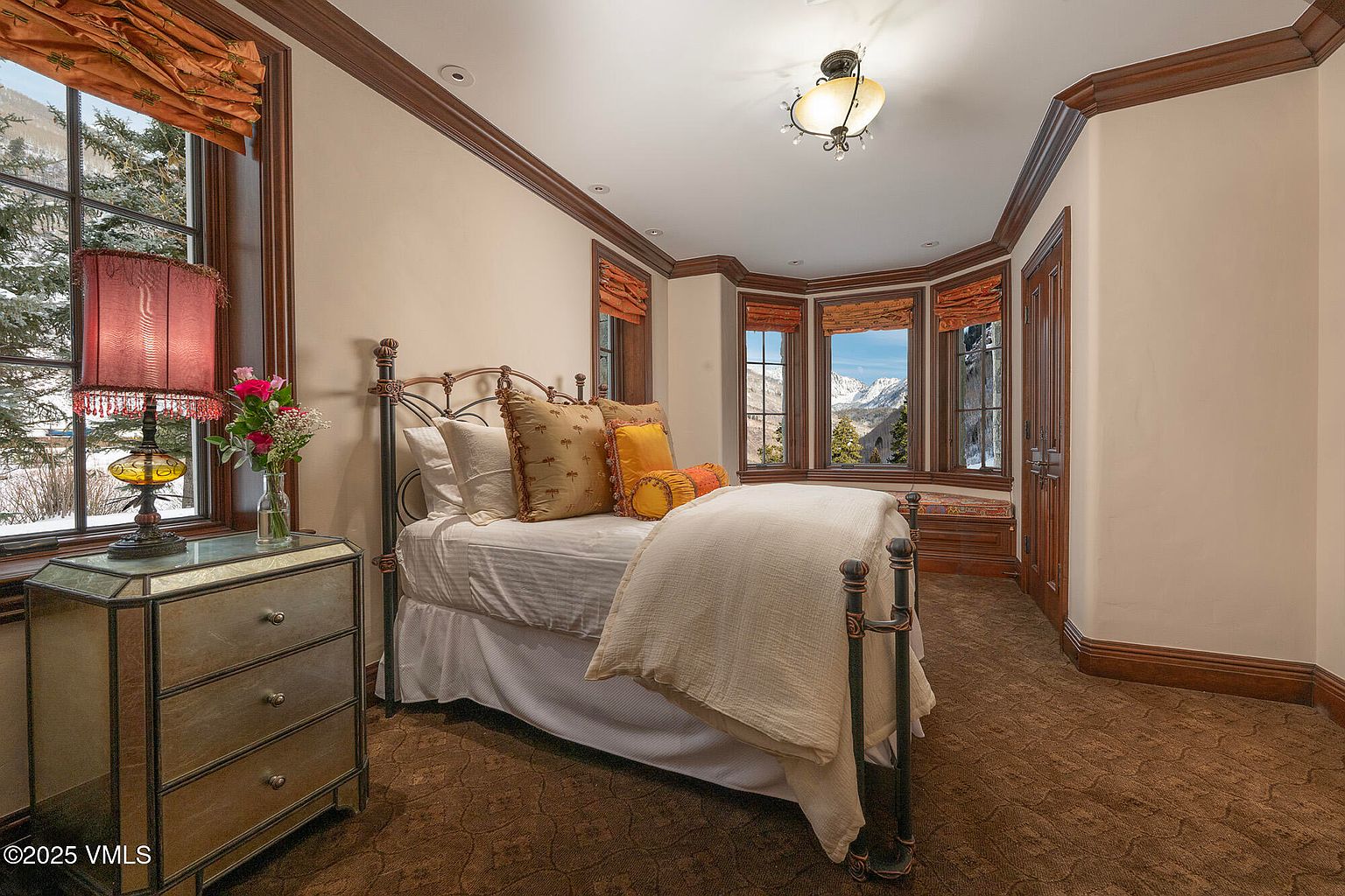 This cozy guest bedroom features a metal-framed bed dressed in neutral bedding, accented by warm-toned throw pillows. A mirrored nightstand with a decorative lamp sits to the left, while a bay window area provides a built-in bench seat with a scenic view of snow-capped mountains. The room is finished with dark wood trim, crown molding, and patterned carpeting, creating a warm and inviting atmosphere.