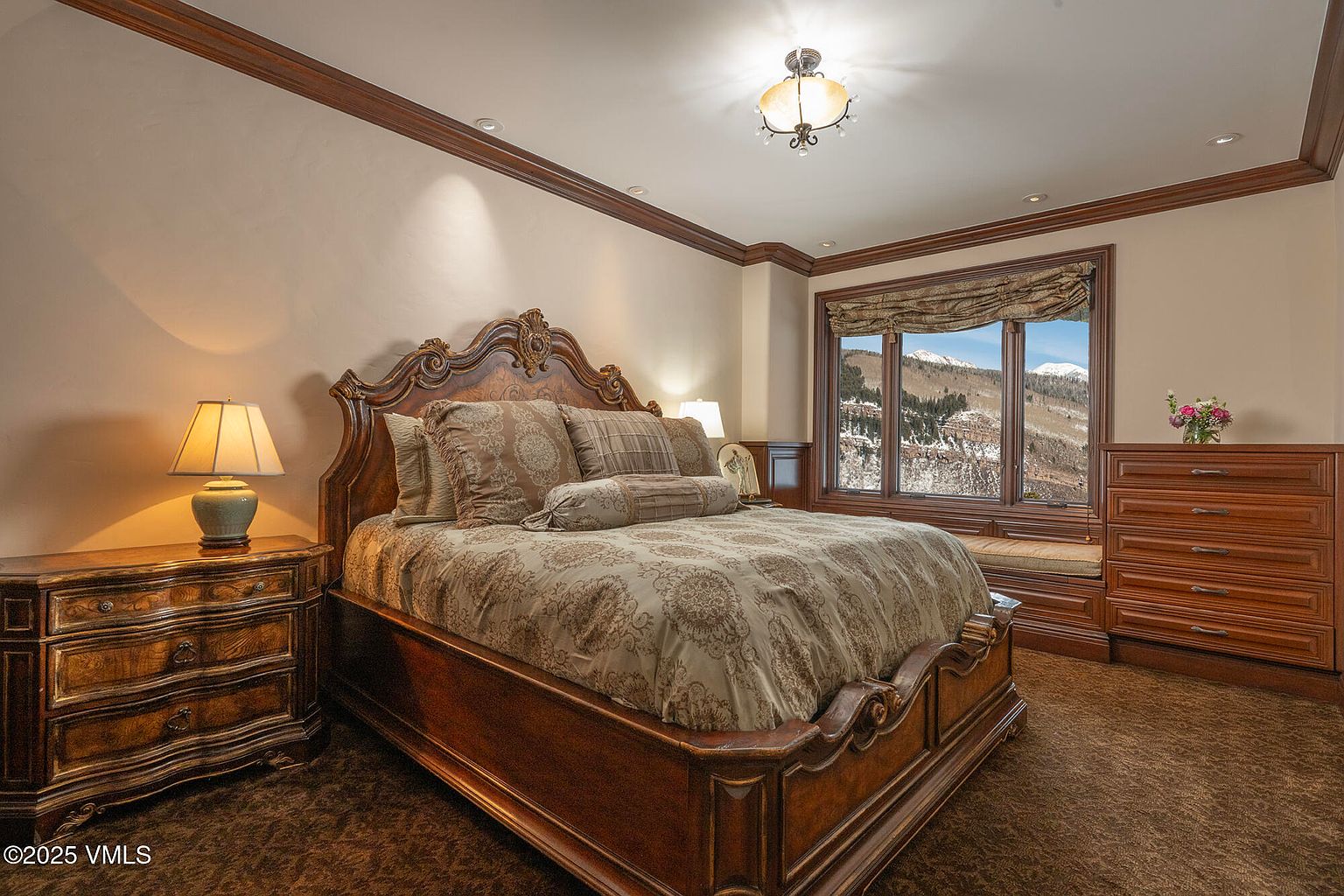 This primary bedroom features a grand, ornate wooden bed frame as the centerpiece, complemented by matching wooden nightstands and a large dresser. The room is decorated in warm, traditional tones with patterned bedding and plush carpeting, while a large window provides a scenic view of snow-covered mountains. A decorative chandelier hangs from the ceiling, adding a touch of elegance to the cozy, well-appointed space.