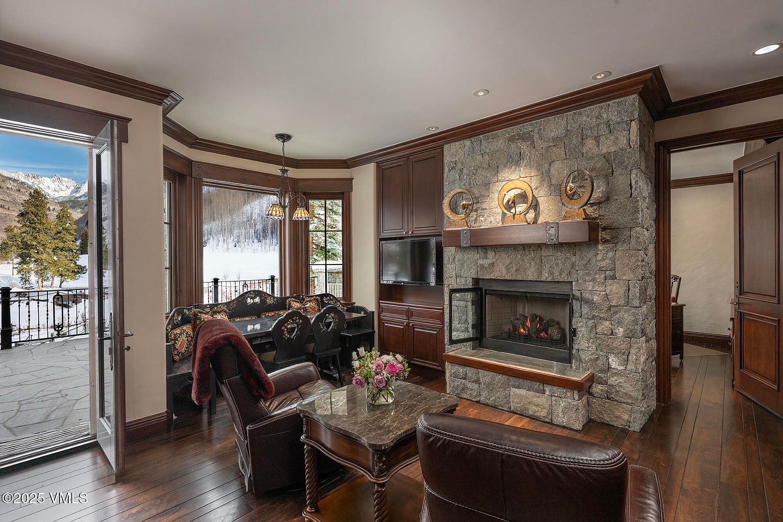 This cozy living area features a prominent stone-clad chimney breast with a built-in fireplace and a wooden mantel topped with decorative circular sculptures. A dark wood-paneled wall houses a television, while a bay window area provides a seating nook with a bench and chairs overlooking a snowy mountain landscape. The room is anchored by leather armchairs and a stone-topped coffee table, creating a warm, rustic atmosphere with direct access to an outdoor stone patio.