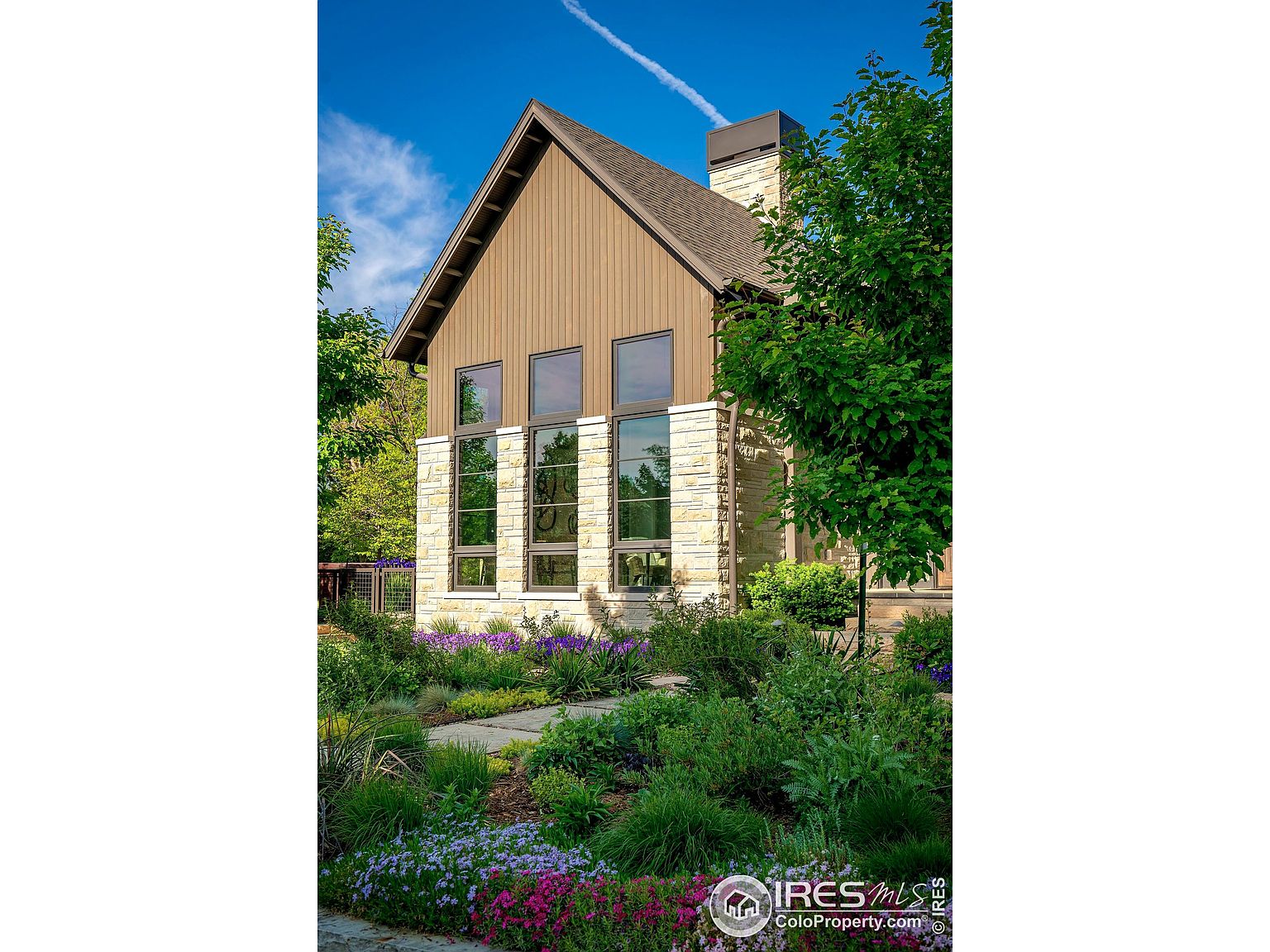 This is a front exterior view of a modern home with a well-maintained garden. The house features a combination of stone and wood siding, with large windows providing ample natural light. The landscaping includes a variety of colorful flowers and lush greenery, enhancing the property's curb appeal.