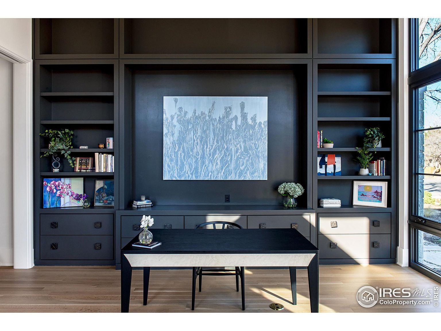 This interior shot showcases a sophisticated home office featuring a built-in dark gray bookcase with open shelving and drawers. A modern desk with a contrasting light-colored top sits in front, complemented by a simple chair. The room is well-lit, highlighting the clean lines and organized space, creating an inviting and productive atmosphere.