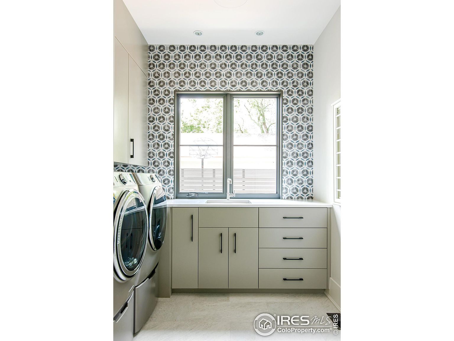 This is a well-organized laundry room featuring a front-loading washer and dryer set, light gray cabinetry with sleek black hardware, and a countertop with a built-in sink. The room is brightened by a window above the countertop, and the wall behind the window is decorated with a geometric patterned wallpaper, adding a touch of style to the functional space.