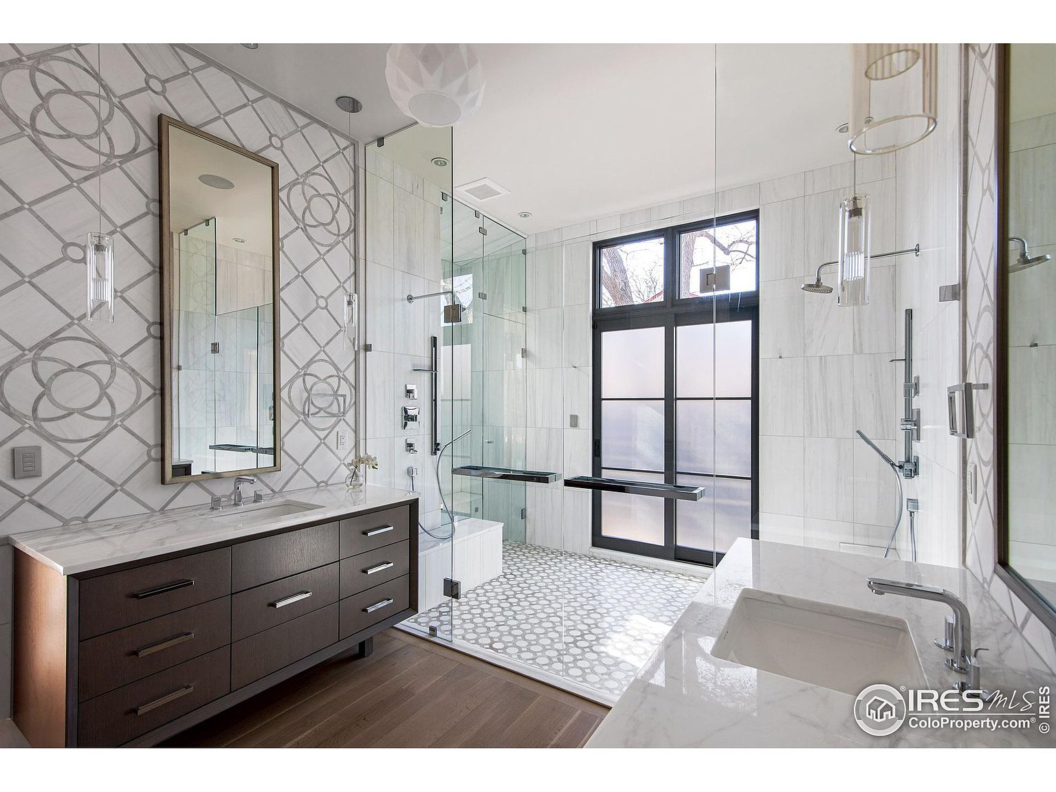 This is a luxurious primary bathroom featuring a double vanity with marble countertops and dark wood cabinetry. A large, glass-enclosed shower with multiple showerheads and a built-in bench is visible, along with a separate toilet area with a frosted glass door. The walls are adorned with decorative tile, and the flooring transitions from wood to patterned tile in the shower area, creating a spa-like atmosphere.