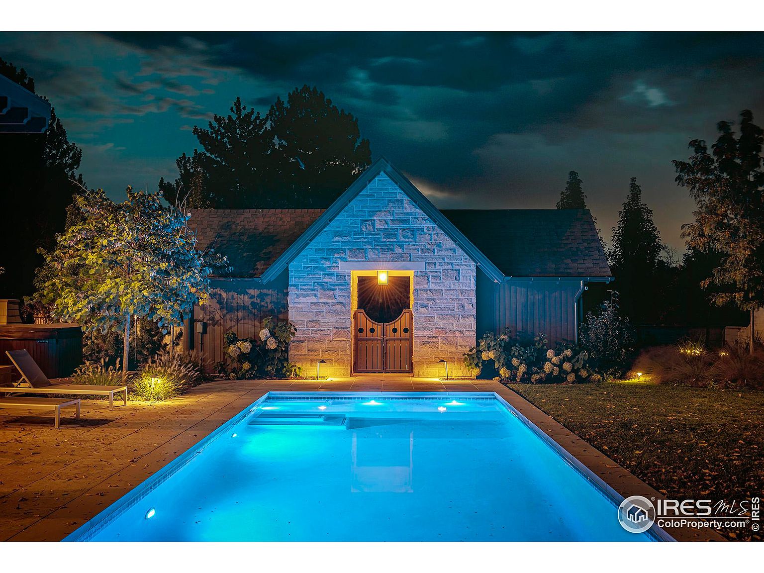 This image showcases a beautifully lit backyard pool area at night. The pool's blue glow contrasts with the warm lighting of the adjacent building, which features a stone facade and wooden doors. The scene is framed by lush landscaping and a clear night sky, creating an inviting and luxurious ambiance.