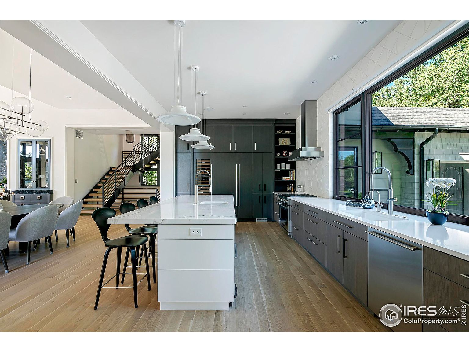 This is a bright and modern kitchen featuring a large island with white countertops and cabinetry, complemented by dark gray cabinets along the wall. Stainless steel appliances and a large window overlooking the exterior add to the contemporary feel. The open layout connects seamlessly to the dining area and staircase, enhancing the sense of spaciousness.
