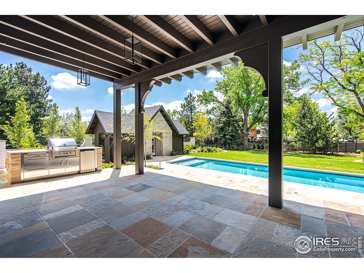 This image showcases a luxurious outdoor patio area with a built-in stainless steel grill and countertop, a stone tile floor, and a covered pergola. A swimming pool is visible in the background, surrounded by a well-manicured lawn and mature trees. The scene evokes a sense of upscale outdoor living and entertaining.