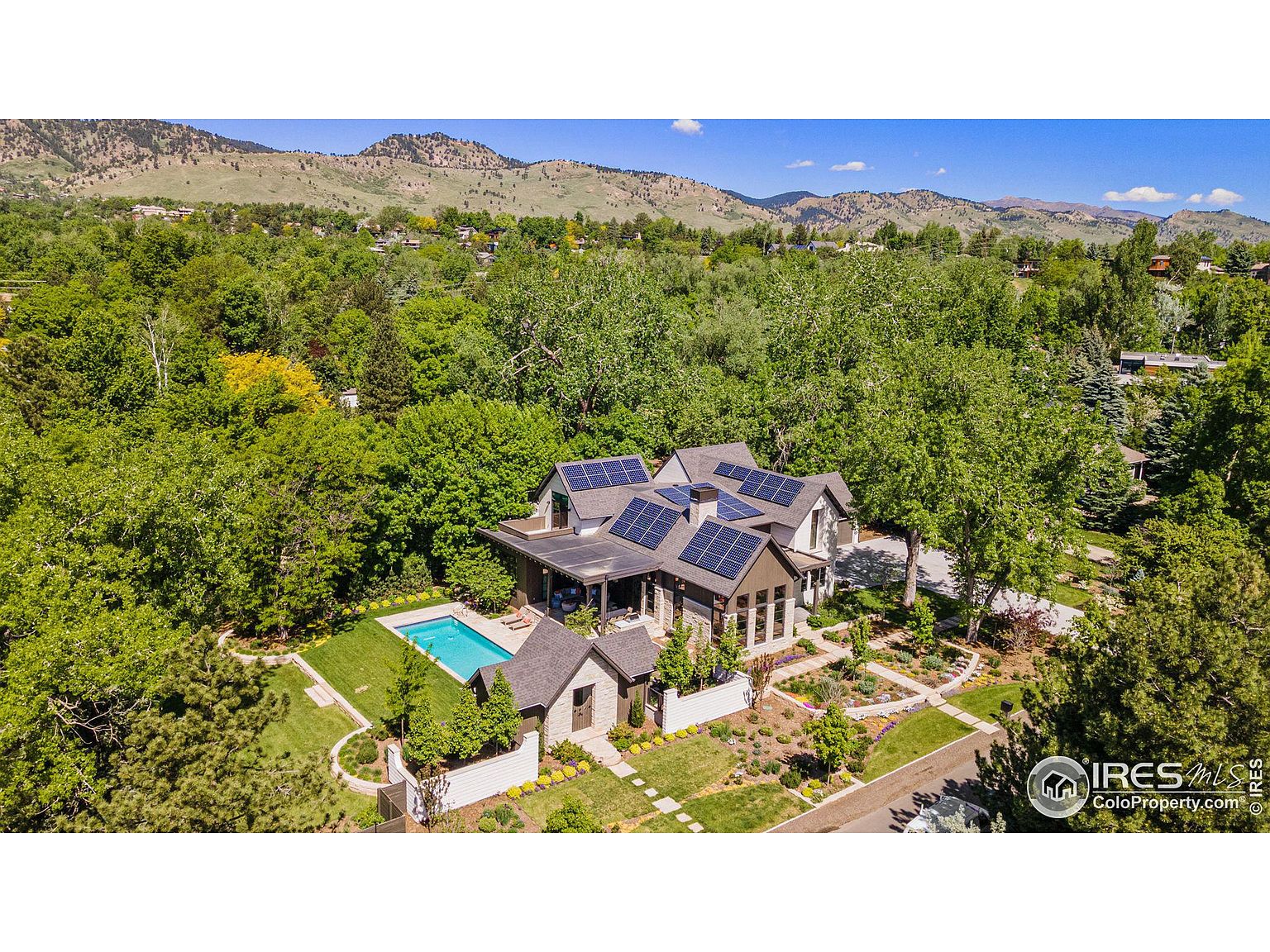 This aerial shot showcases a luxurious home nestled among lush greenery, with mountains visible in the background. The property features a swimming pool, well-manicured lawns, and a charming garden. Solar panels are installed on the roof, highlighting the home's energy efficiency.