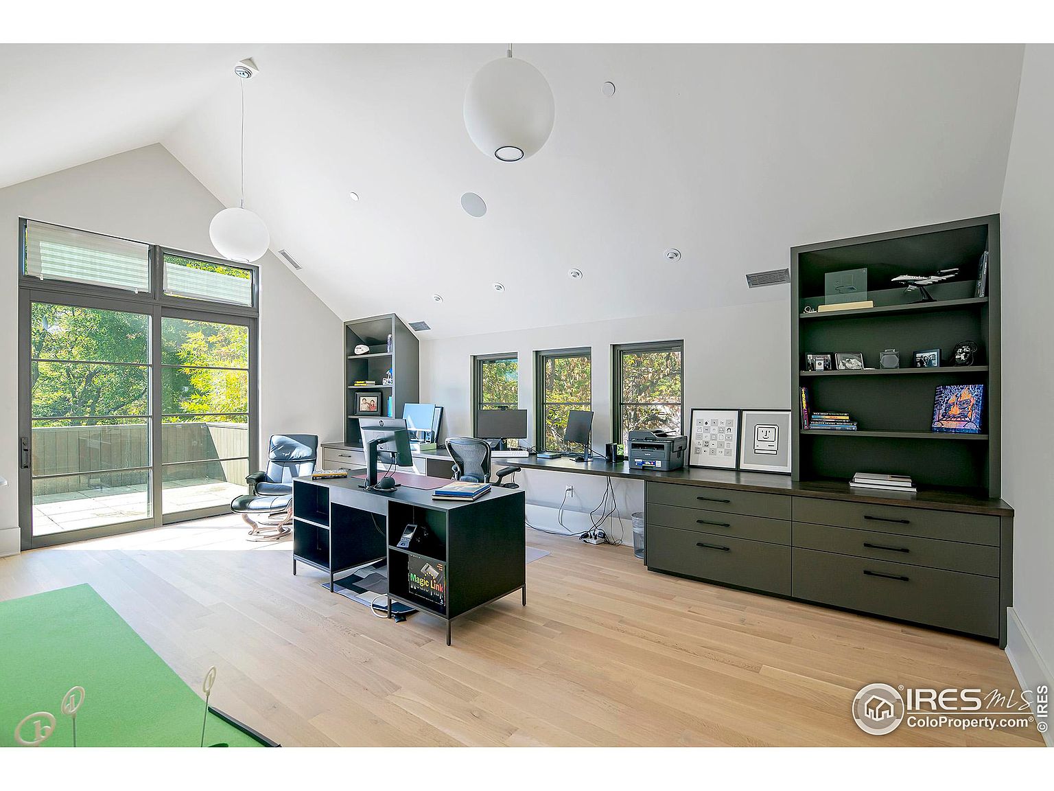This is an interior shot of a modern office space featuring a large desk, built-in shelving, and a wall of windows providing natural light. The room has light wood flooring and a neutral color palette, creating a bright and airy atmosphere. A putting green is visible in the foreground, adding a unique touch to the workspace.