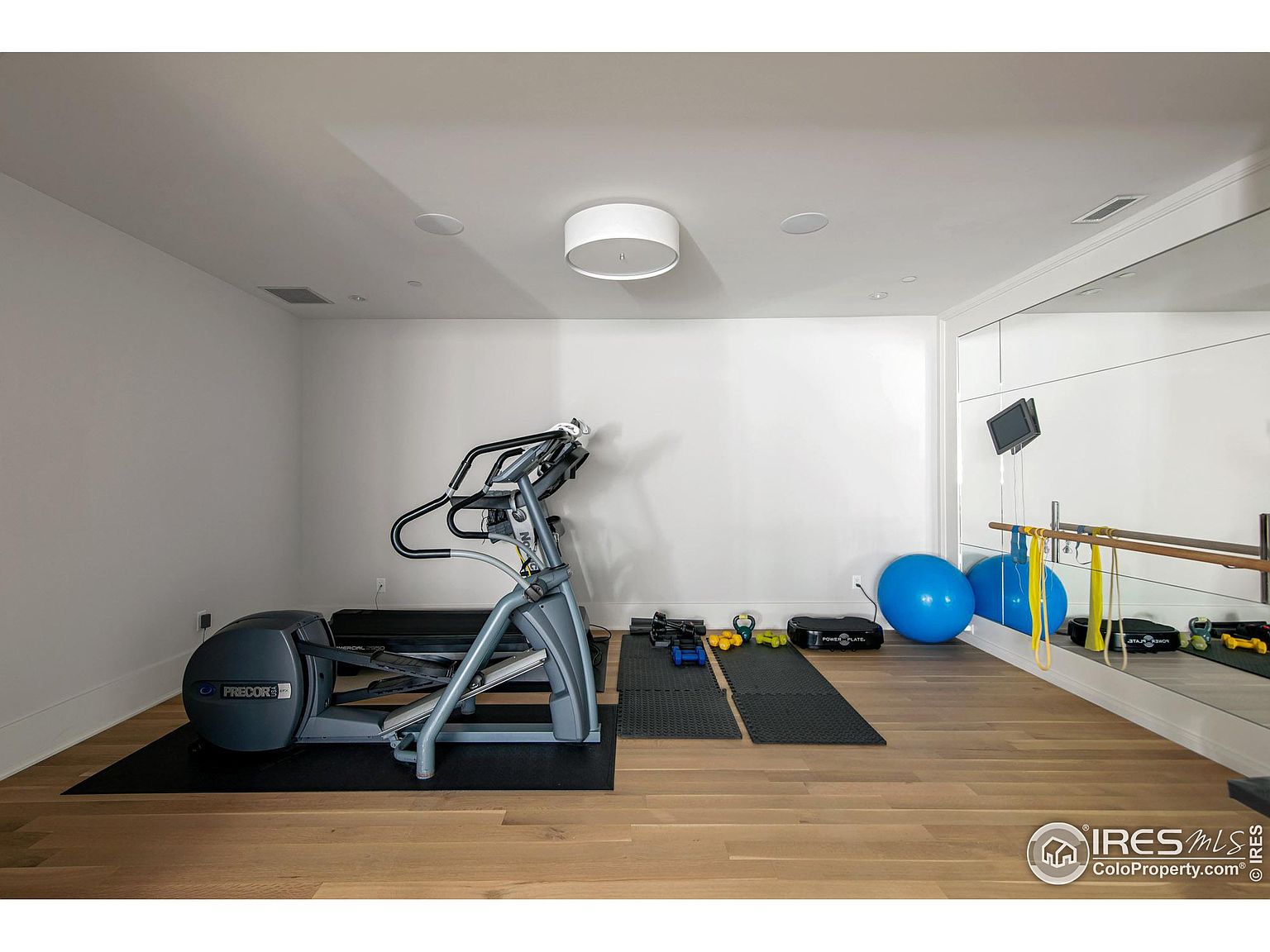 This is an interior shot of a home gym featuring an elliptical machine on a black mat, various weights and exercise equipment, and a mirrored wall with a ballet barre. The room has hardwood floors, white walls, and recessed lighting, creating a clean and functional workout space. The perspective is from a medium distance, showcasing the layout and equipment.