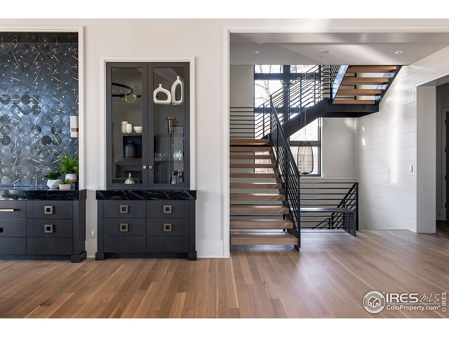 This interior shot showcases a modern hallway with a striking staircase. The staircase features wooden steps and a black metal railing, creating a contemporary aesthetic. Adjacent to the stairs is a built-in cabinet with glass doors and a dark countertop, adding a touch of elegance to the space.