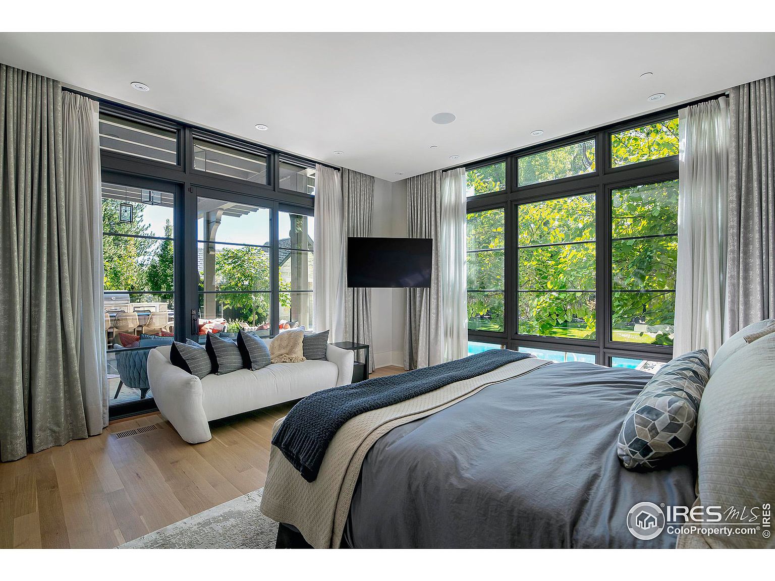 This is a primary bedroom featuring a large bed with gray bedding, a white sofa, and floor-to-ceiling windows offering views of the exterior. The room is decorated in neutral tones with gray curtains and hardwood floors, creating a serene and luxurious atmosphere. A flat-screen TV is mounted on the wall.