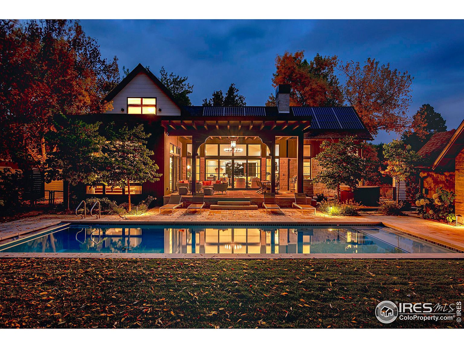 This image showcases the rear exterior of a luxurious home, highlighting a stunning pool area at dusk. The pool reflects the warm light emanating from the house, which features a covered patio with comfortable seating. Mature trees surround the property, adding to the sense of privacy and tranquility, while solar panels are visible on the roof.