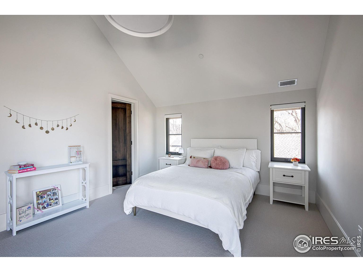 This is a bright and airy bedroom featuring a white bed with matching nightstands, complemented by neutral-toned walls and carpet. The room includes two windows that provide natural light, and a decorative console table adds a touch of style. The overall impression is clean, comfortable, and inviting.