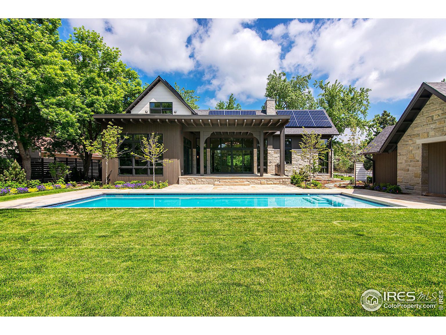 This image showcases a stunning backyard featuring a modern home with solar panels, a pristine swimming pool, and a lush green lawn. The architectural style blends natural materials with contemporary design, creating an inviting outdoor living space. The perspective is a wide shot, emphasizing the spaciousness and luxurious feel of the property.