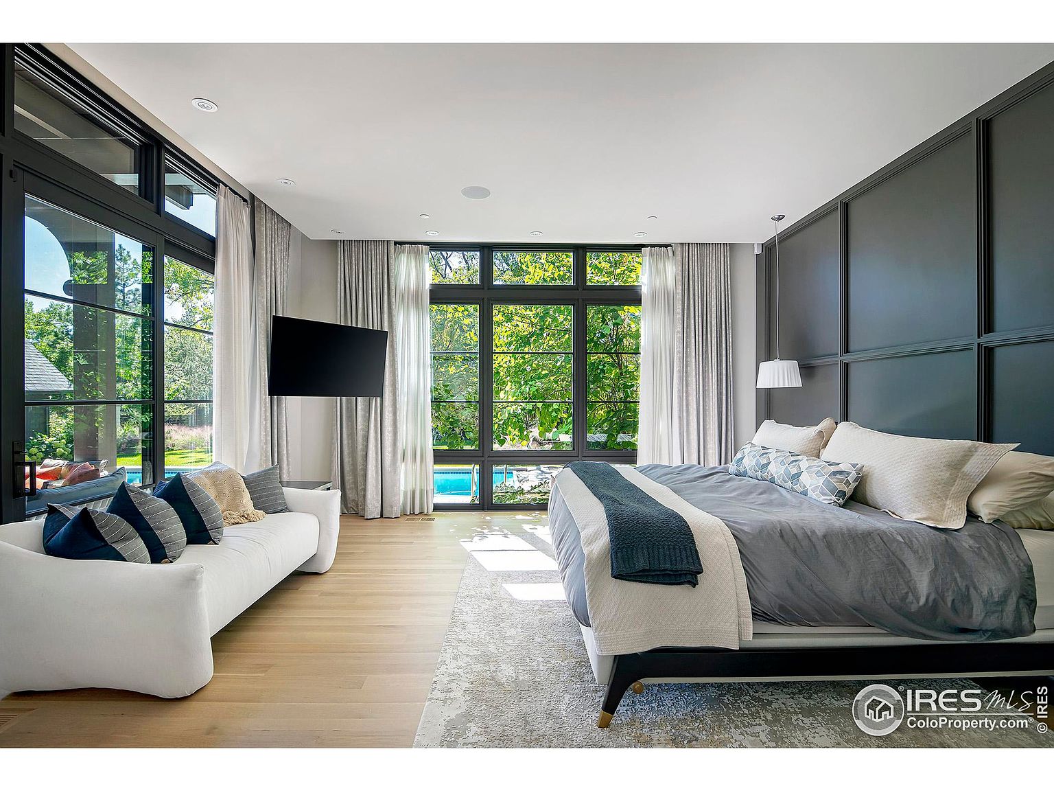 This is a luxurious primary bedroom featuring a large bed with a gray duvet and decorative pillows, a white sofa, and a wall-mounted television. Large windows offer views of the exterior greenery and pool, while elegant curtains frame the windows. The room is well-lit with natural light and features a modern, sophisticated design.