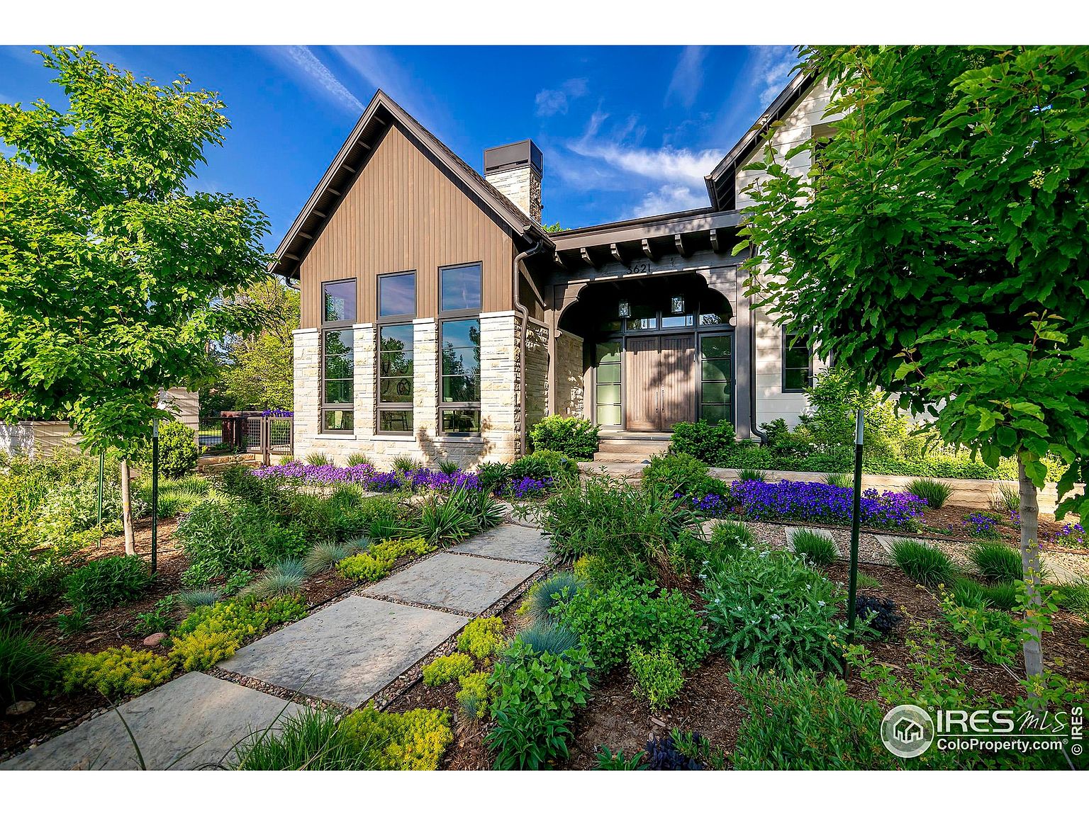 This is a front exterior view of a beautifully landscaped home. The house features a combination of stone and wood siding, with large windows and a covered entryway. The front yard is meticulously maintained with a variety of plants and a stone pathway leading to the front door, creating an inviting and upscale curb appeal.
