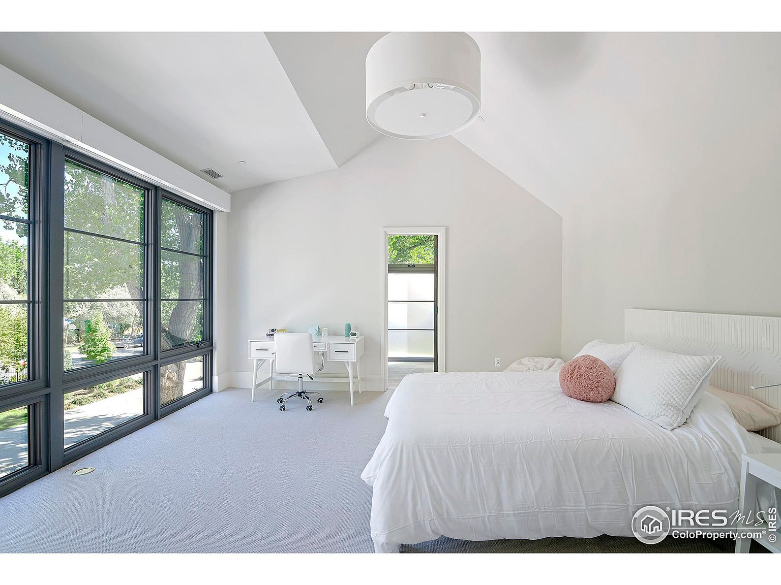 This is a bright and airy primary bedroom featuring large windows that offer ample natural light and a view of the outdoors. The room is decorated in a minimalist style with white walls, light carpet, and a simple white bed. A small desk and chair are positioned near the windows, creating a functional workspace.