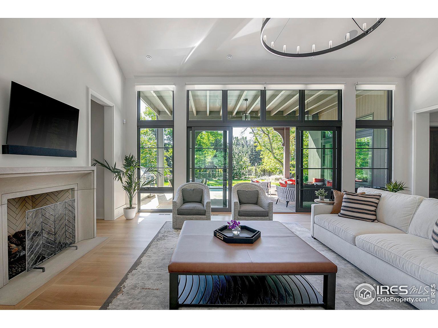 This is a bright and airy living room featuring a modern design with large windows that provide ample natural light and a view of the outdoor space. The room includes a fireplace, a large flat-screen TV, comfortable seating, and a stylish coffee table. The overall impression is one of sophistication and comfort, ideal for relaxation and entertaining.