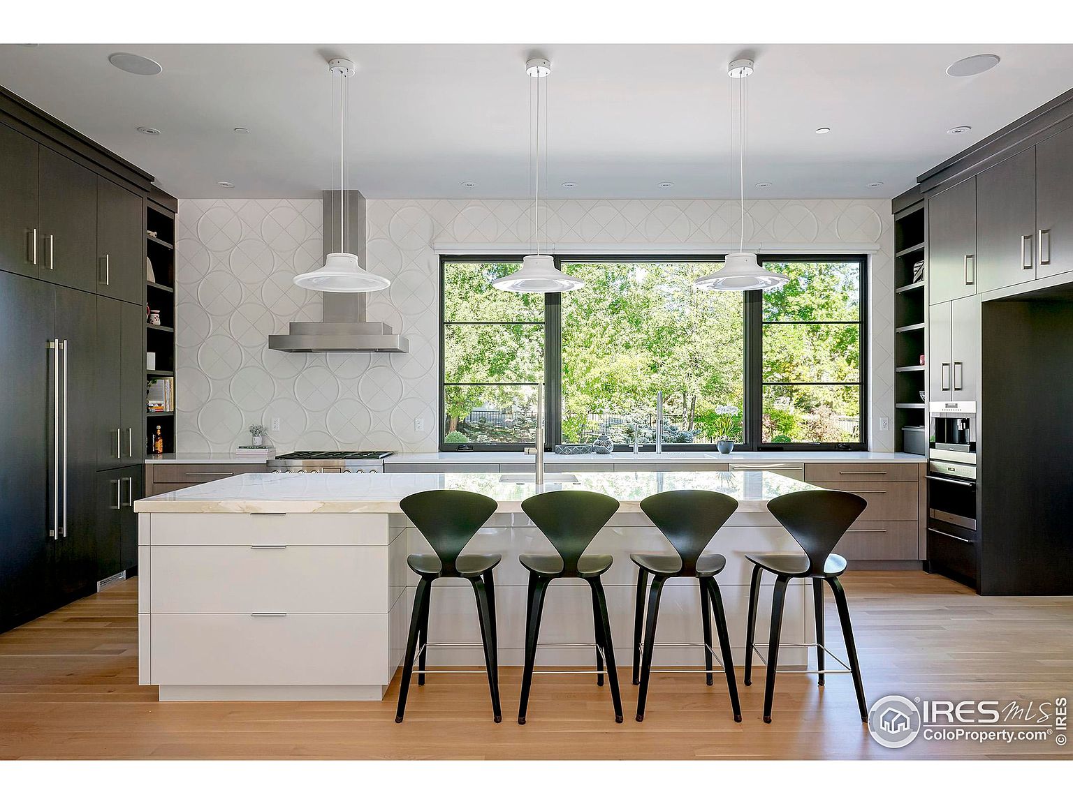 This is a well-lit, modern kitchen featuring a large island with a white countertop and seating for four. The kitchen includes dark cabinetry, stainless steel appliances, and a large window providing natural light and a view of the outdoors. The overall design is sleek and contemporary, creating a welcoming and functional space.