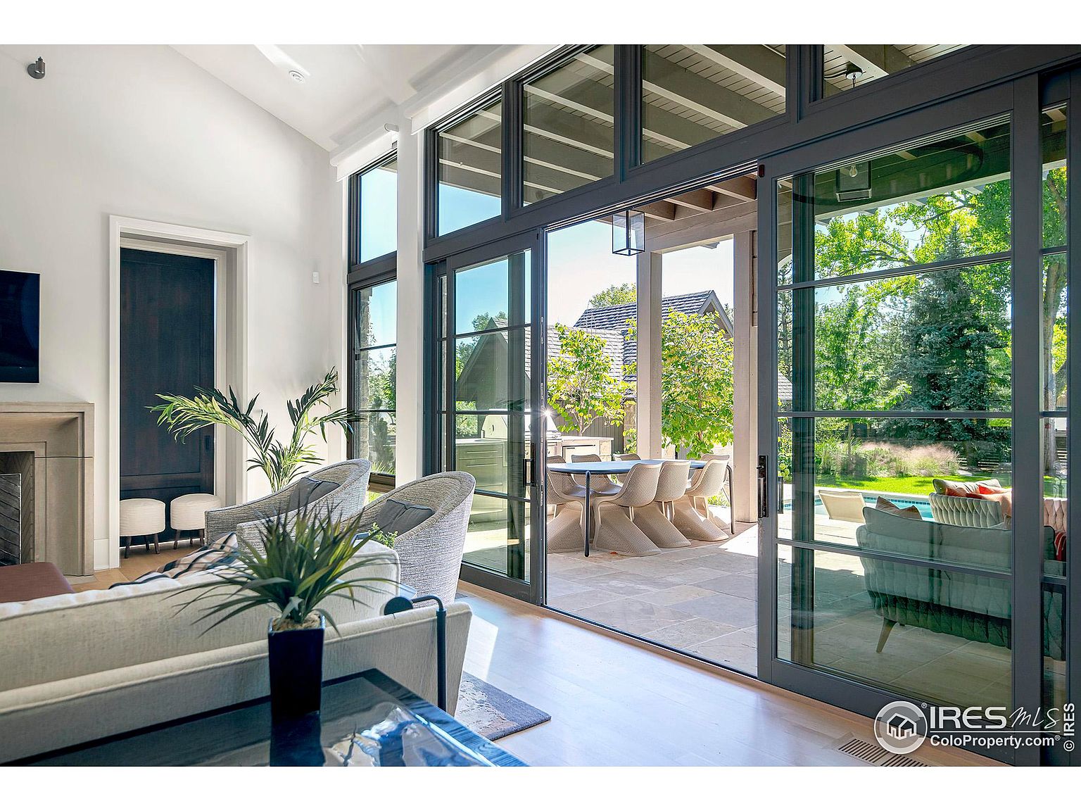 This image showcases a bright and airy living room that seamlessly blends indoor and outdoor living. Large sliding glass doors open to a patio with an outdoor dining area and a view of the pool and backyard. The interior features high ceilings, modern furniture, and stylish decor, creating an inviting and luxurious atmosphere.