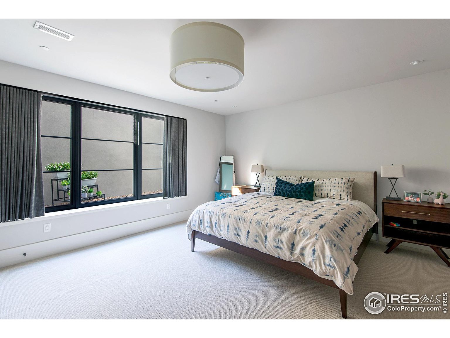 This is a well-lit primary bedroom featuring a large window with dark frames and gray curtains, offering a view of an outdoor planter. The room is furnished with a bed dressed in a patterned duvet, two nightstands with lamps, and a full-length mirror. The overall aesthetic is modern and minimalist, with a neutral color palette and clean lines.