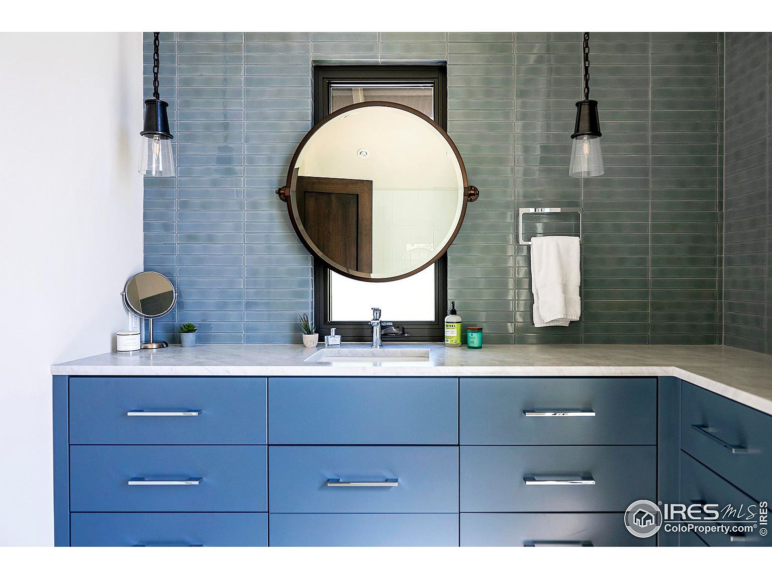 This is a well-lit bathroom featuring a blue vanity with modern silver hardware and a white countertop. The backsplash is a light blue tile, and a round mirror hangs above the sink. Pendant lights flank the mirror, adding a touch of elegance to the space.