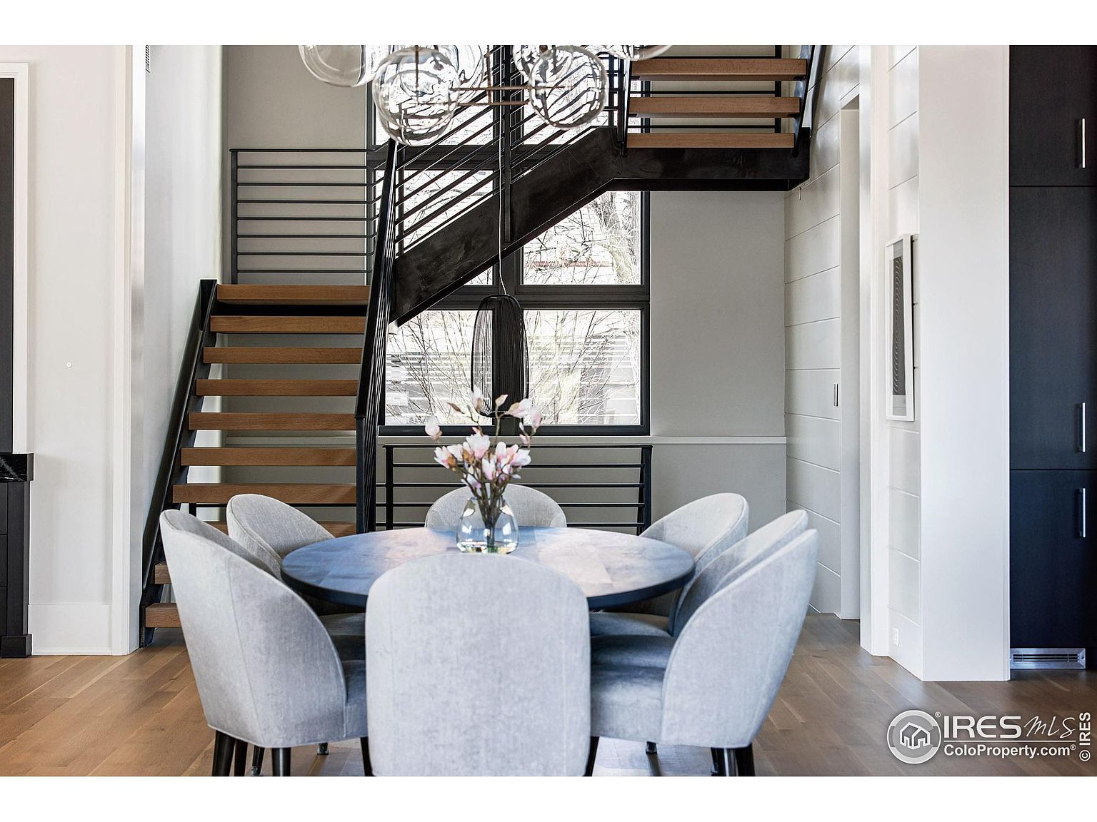 This is an interior shot of a dining room featuring a round table with six light gray upholstered chairs. A modern staircase with wooden steps and black metal railings is visible in the background, along with a window providing natural light. The room has hardwood floors and a contemporary design, creating an inviting and stylish dining space.