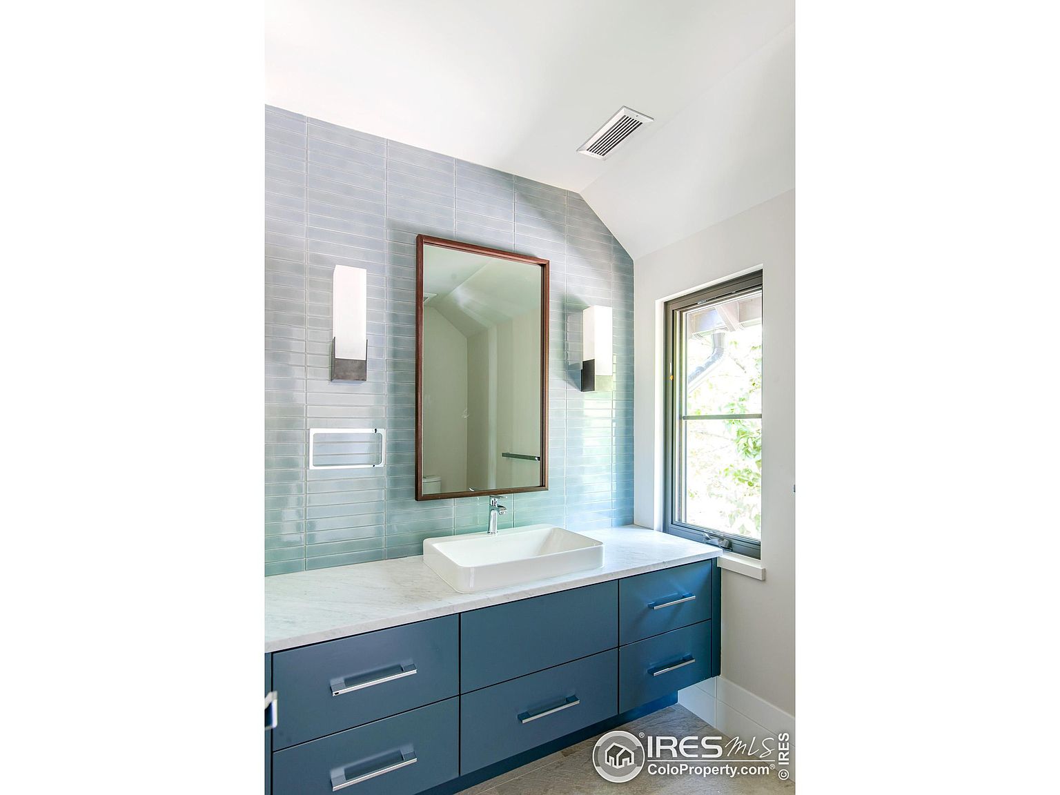 This is a well-lit bathroom featuring a blue vanity with a white countertop and a rectangular vessel sink. The wall is tiled in a light blue, horizontal pattern, complemented by a framed mirror and modern sconces. A window provides natural light, enhancing the clean and contemporary aesthetic of the space.